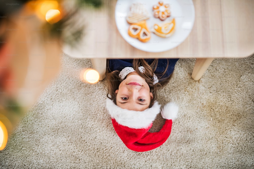 Una vista dall'alto di una bambina con il cappello di Babbo Natale sdraiata sul pavimento sotto un tavolo nel periodo natalizio.