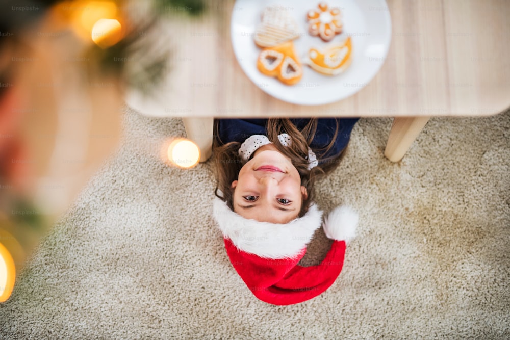 Eine Draufsicht auf ein kleines Mädchen mit Weihnachtsmütze, das zur Weihnachtszeit unter einem Tisch auf dem Boden liegt.