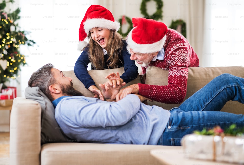 Ein kleines Mädchen und ihr Großvater mit Weihnachtsmütze wecken ihren Vater, der zur Weihnachtszeit auf einem Sofa schläft.