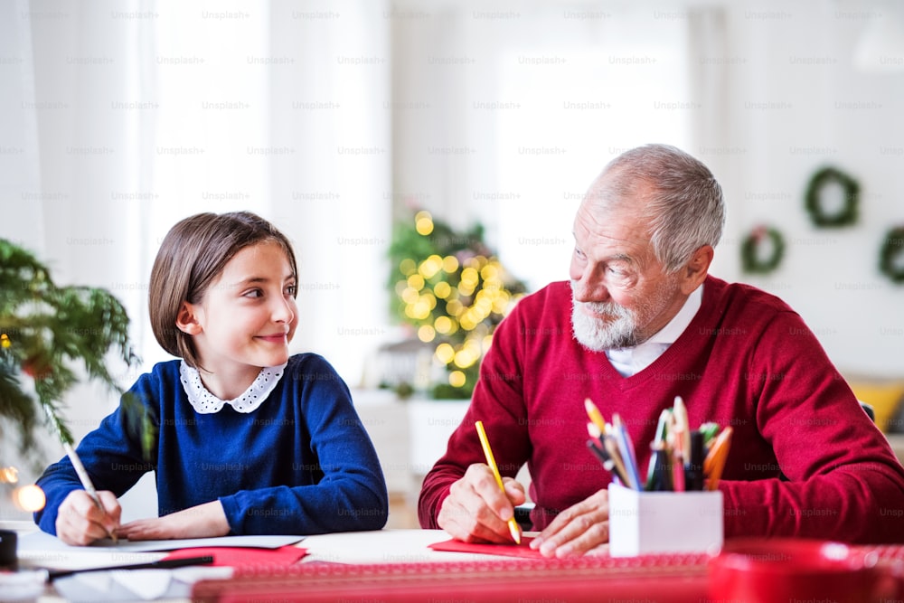 Ein kleines Mädchen und ihr Großvater sitzen an einem Tisch und schreiben gemeinsam Weihnachtskarten.