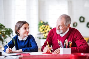 A small girl and her grandfather sitting at a table, writing Christmas cards together.