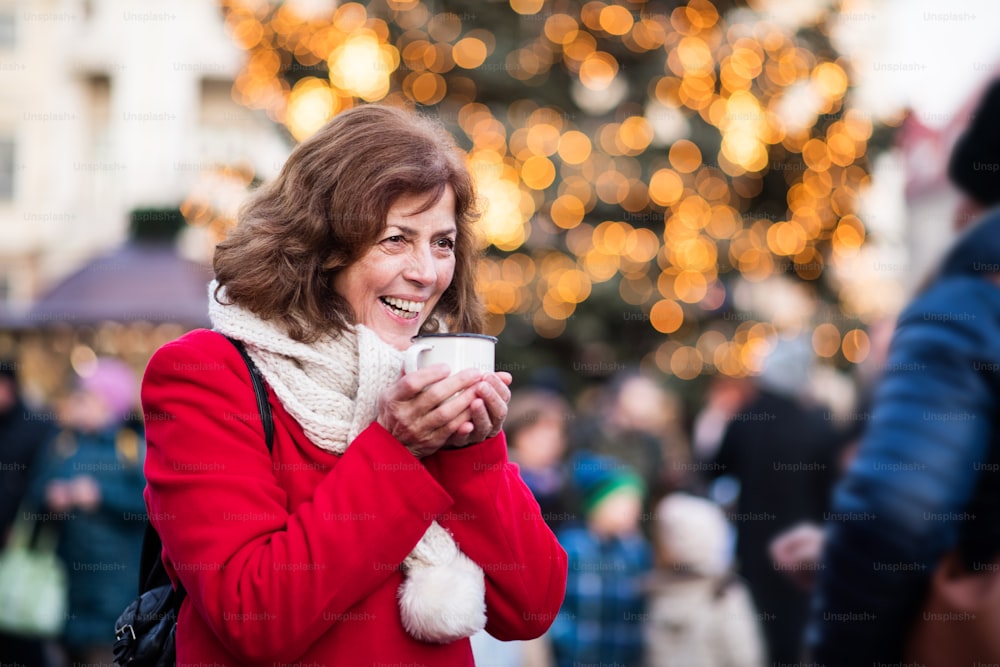 Feliz mujer mayor en un mercado navideño al aire libre, sosteniendo una taza esmaltada. Horario de invierno.