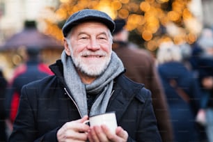 Homem sênior feliz em um mercado de Natal ao ar livre, segurando copo esmaltado. Horário de inverno.