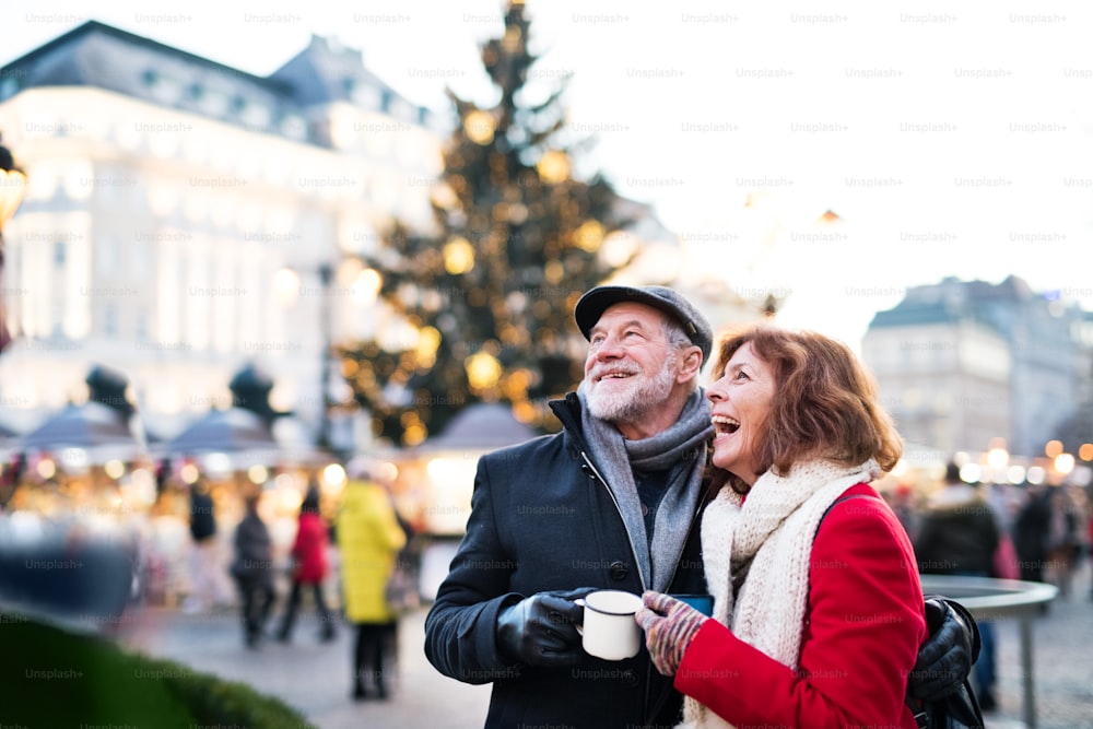 屋外のクリスマスマーケットで、エナメルを塗ったカップを持つ幸せな老夫婦。冬時間。
