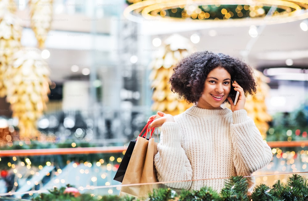 Un retrato de una adolescente con un teléfono inteligente y bolsas de papel en un centro comercial en Navidad, haciendo una llamada telefónica. Espacio de copia.