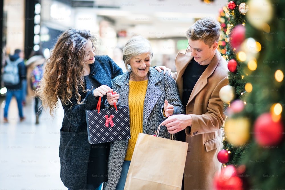 Un ritratto di nonna anziana e nipoti adolescenti con sacchetti di carta in piedi nel centro commerciale nel periodo natalizio.
