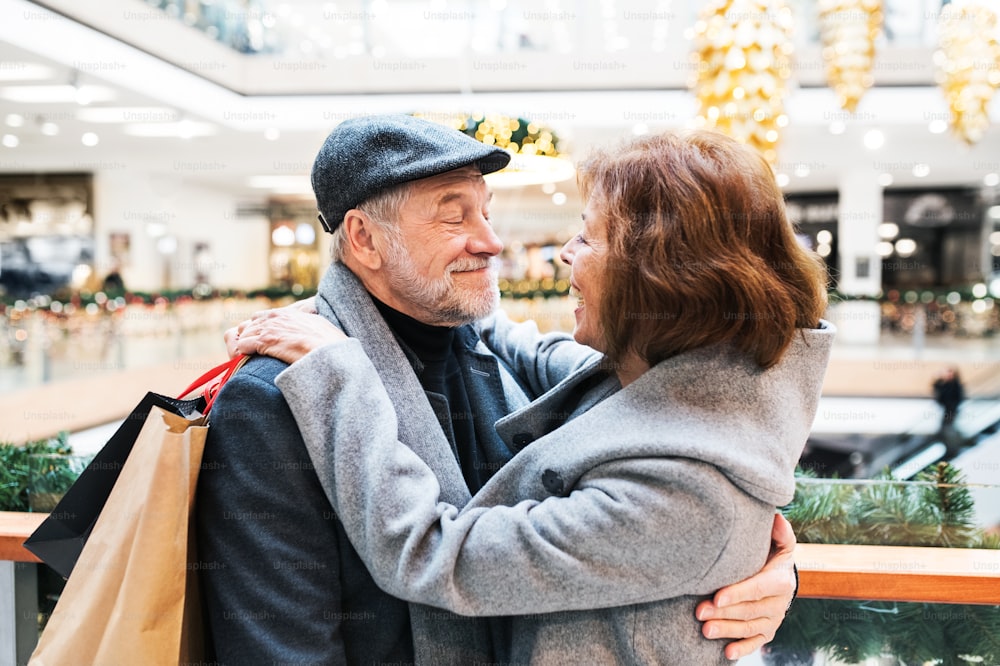 Casal idoso feliz com sacos de papel no centro comercial olhando um para o outro, abraçando-se.