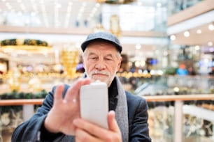 Uomo anziano che fa shopping natalizio. Uomo che scatta selfie con uno smartphone. Centro commerciale nel periodo natalizio.