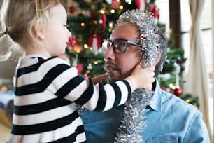 Junger Vater mit seiner niedlichen kleinen Tochter, die den Weihnachtsbaum schmückt. Mädchen, das ihm silberne Lametta-Girlande um den Kopf gibt.
