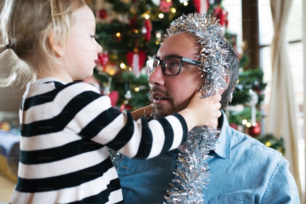 Young father with his cute little daughter decorating Christmas tree. Girl giving him silver tinsel garland around his head.