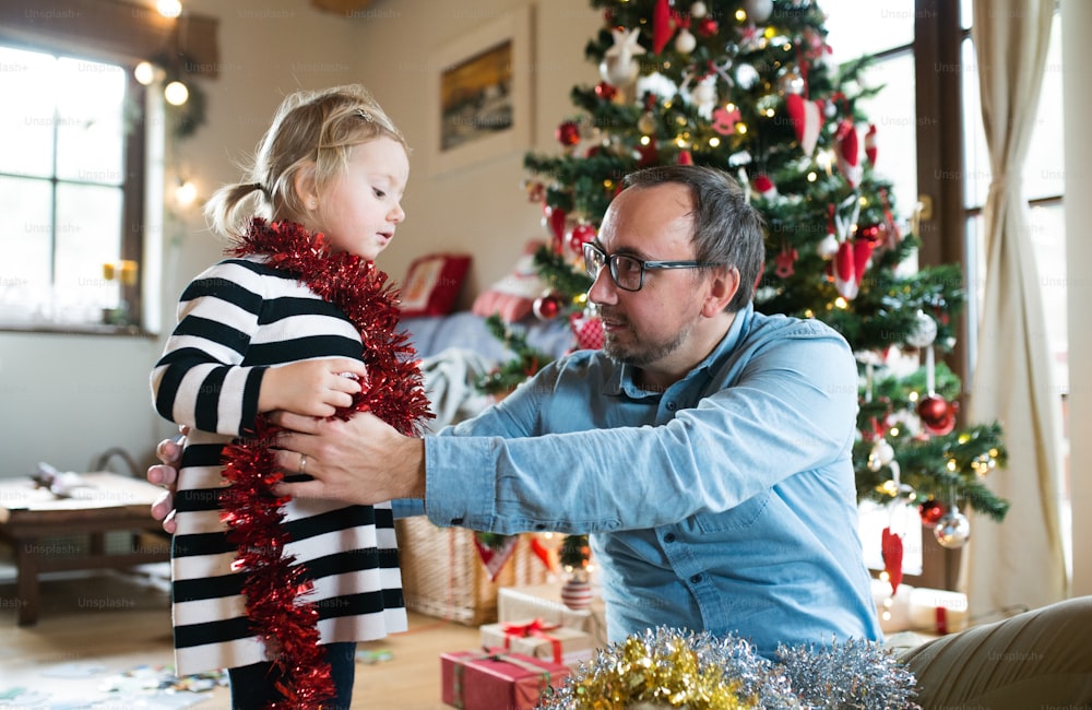 Young father with his cute little daughter decorating Christmas tree. Man wrapping red tinsel garland around the girl.