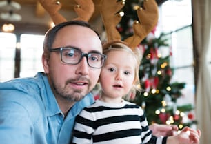 Padre joven con su linda hijita en el árbol de Navidad, vistiendo astas de reno marrón, tomándose selfie.
