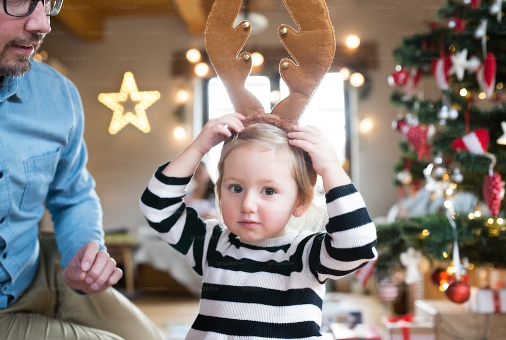 Young father with his cute little daughter at the Christmas tree, wearing brown reindeer antlers.
