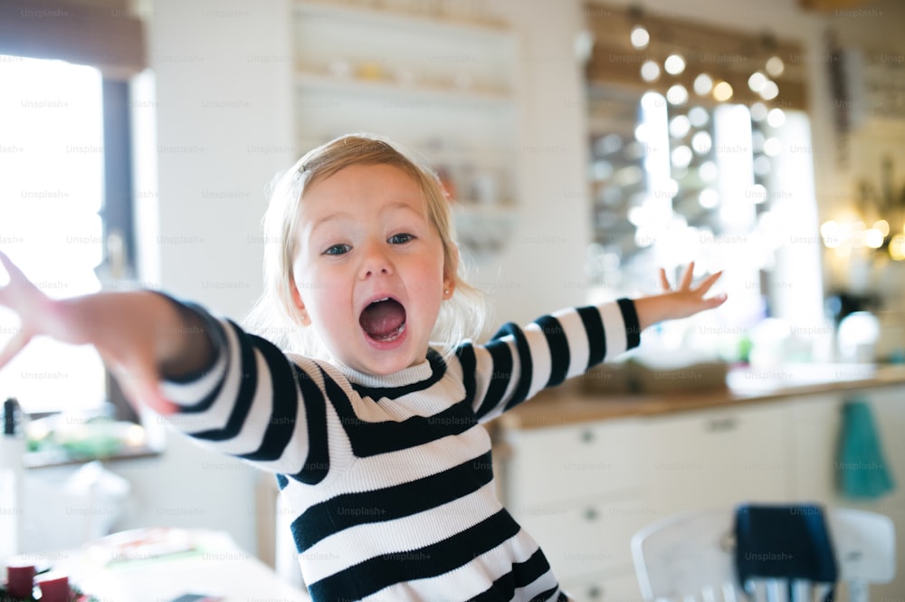 Cute little blond girl in striped dress sitting on kitchen table, shouting, arms stretched. Christmas season.