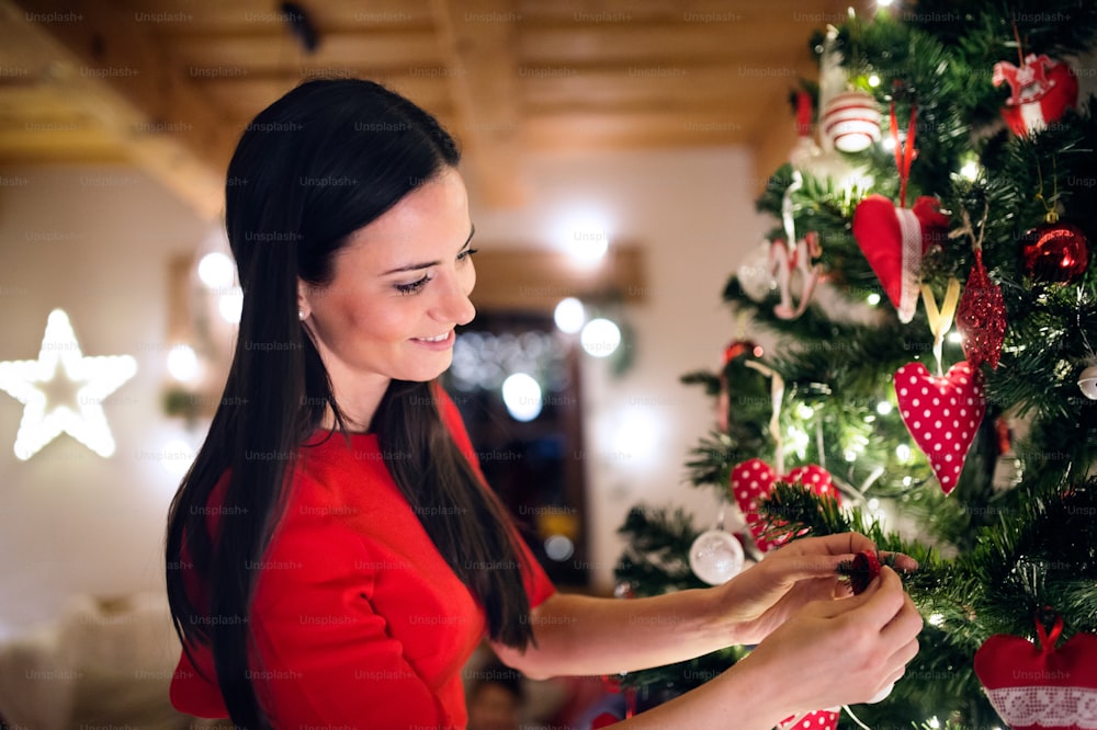 Beautiful young woman in red dress in front of illuminated Christmas tree inside in her house decorating it.