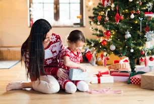 Beautiful young mother with little daughter at Christmas tree at home unpacking presents.