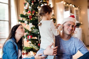 Beautiful young family with little girl decorating Christmas tree at home. Daughter giving father Christmas ornaments on his ears.
