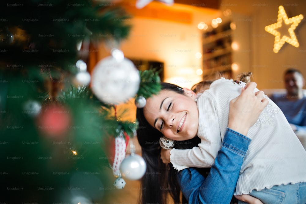 Beautiful young mother with little daughter at home decorating Christmas tree together, hugging.