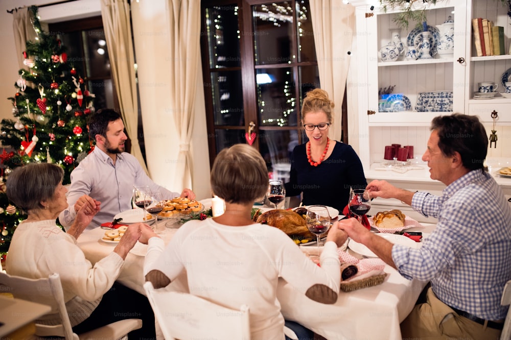 Linda família grande sentada à mesa, celebrando Christmat juntos em casa, de mãos dadas, orando. Árvore de Natal iluminada atrás deles.