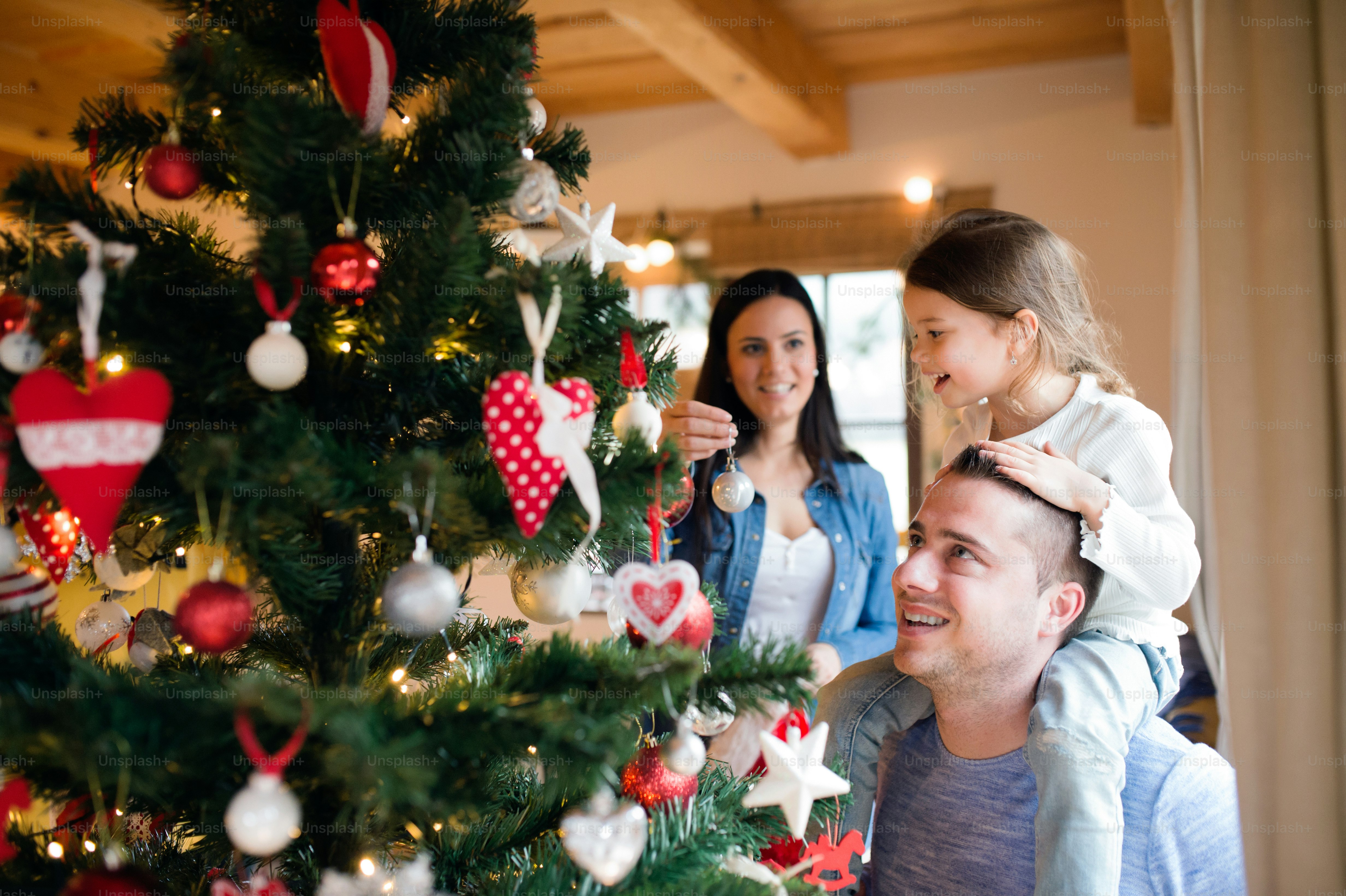 Beautiful young family with little girl decorating Christmas tree at home. Father giving daughter piggyback.