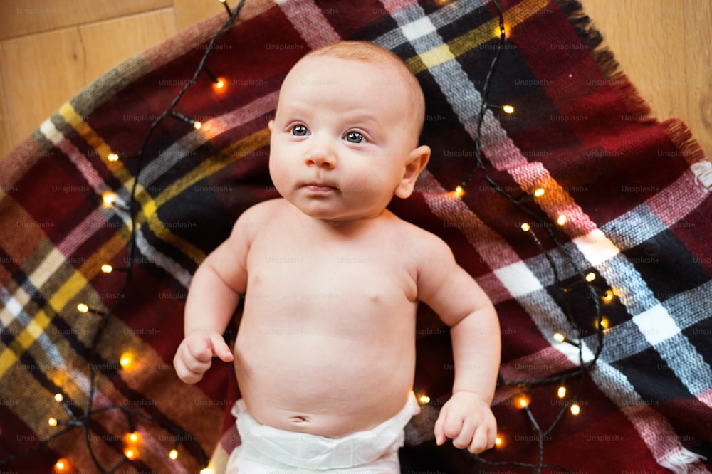 크리스마스 트리 아래 귀여운 아기가 체크무늬 담요에 누워 있다. 높은 각도 보기.