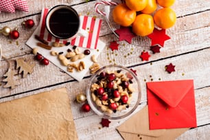 Christmas composition. Bowl with dried fruit, cranberries and nuts. Cup of coffee. Tangerines on plate. Christmas cards and decorations. Various objects laid on table. Studio shot, wooden background. Copy space.