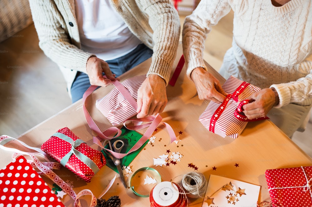 Irreconocible pareja mayor con suéteres blancos de lana sentados a la mesa envolviendo regalos de Navidad juntos. Vista aérea.