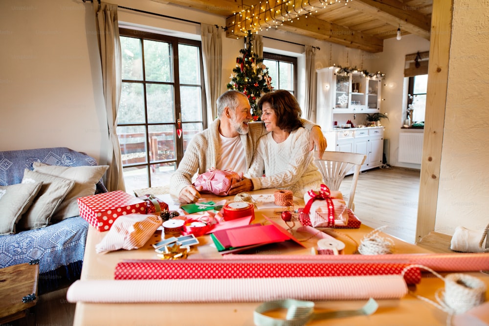 Beautiful senior couple in white woolen sweaters sitting at the table wrapping Christmas gifts together.