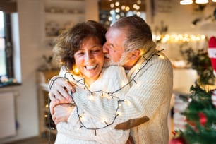 Beautiful senior couple in white woolen sweaters hugging at home. Man and woman having fun at Christmas time.