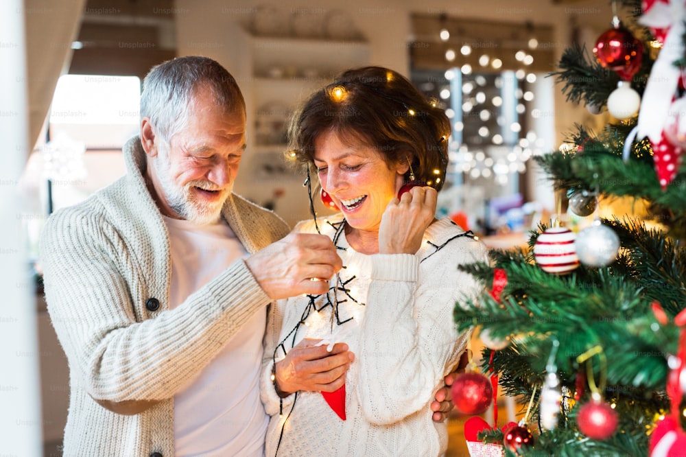 Beautiful senior couple in white woolen sweaters decorating Christmas tree inside their house. Man and woman having fun.