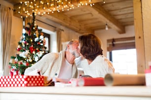Beautiful couple in white woolen sweaters sitting at the table wrapping Christmas gifts together. Senior couple kissing.