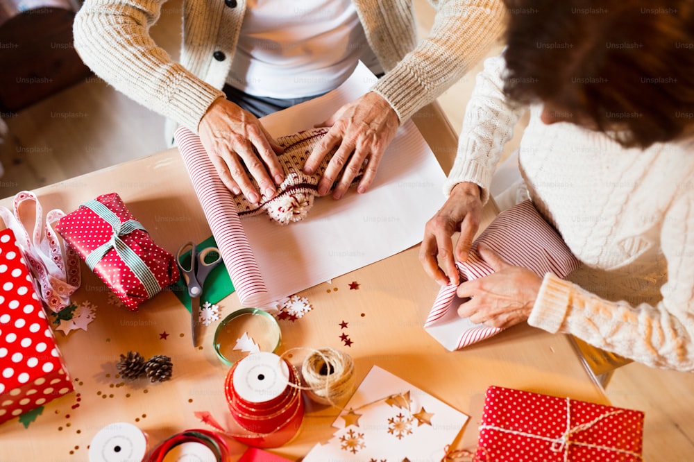 Coppia anziana irriconoscibile in maglioni di lana bianca seduti al tavolo che avvolgono insieme i regali di Natale. Veduta aerea.
