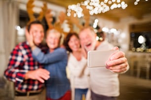 Senior friends wearing reindeer antlers headbands at Christmas time, having fun. Two men and two women with smartphone taking selfie.