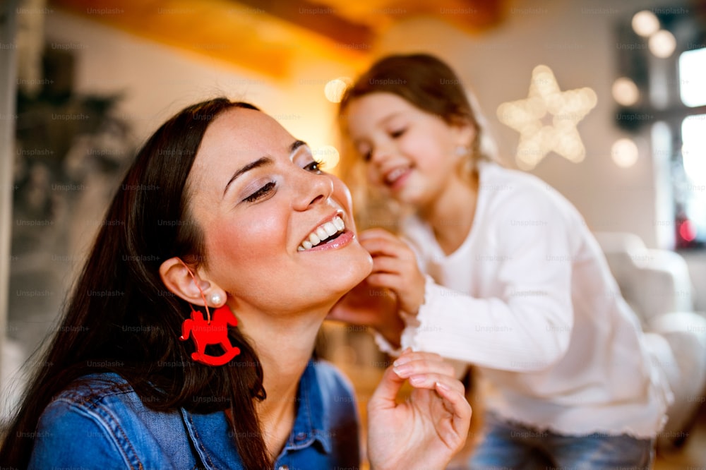 Schöne junge Mutter mit kleiner Tochter zu Hause, die gemeinsam den Weihnachtsbaum schmückt. Kleines Mädchen gibt ihrer Mutter Weihnachtsschmuck auf den Ohren.