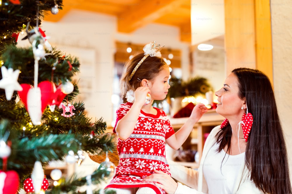Beautiful young mother with little daughter at Christmas tree at home, putting Christmas ornaments on ears.