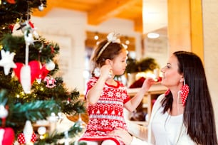 Beautiful young mother with little daughter at Christmas tree at home, putting Christmas ornaments on ears.