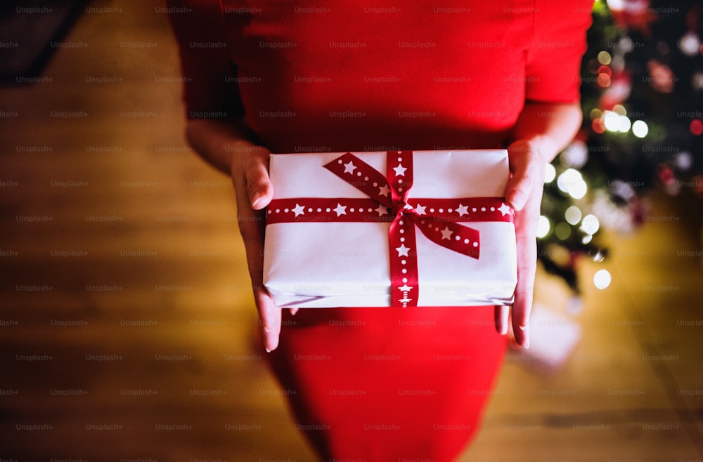 Unrecognizable young woman in red dress in front of illuminated Christmas tree inside in her house holding Christmas present.