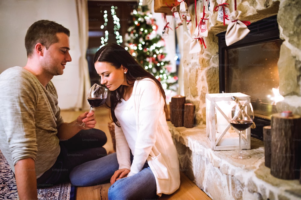 Bella giovane coppia seduta sul pavimento davanti al camino, bevendo vino rosso. Tempo di Natale.