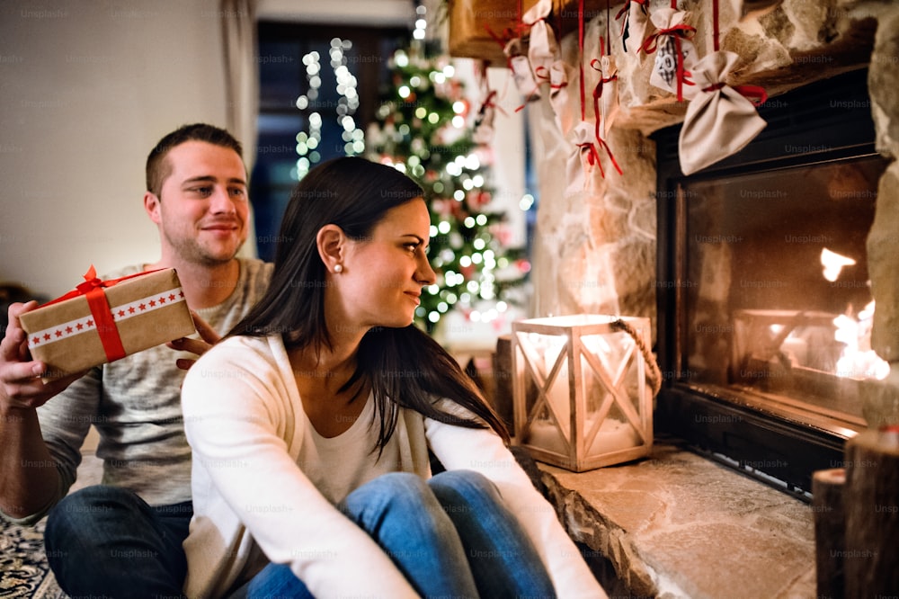 Beau jeune couple assis par terre devant la cheminée. Homme offrant un cadeau de Noël à sa belle femme.