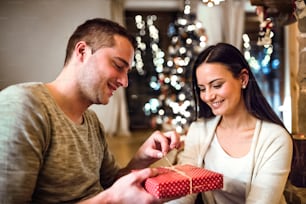 Beautiful young couple sitting on the floor in front of fireplace. Man adn woman giving presents to each other.