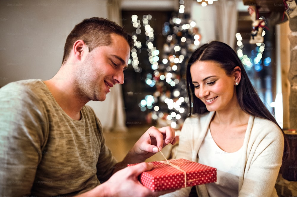 Beau jeune couple assis par terre devant la cheminée. L’homme et la femme se donnent des cadeaux.