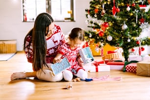 Hermosa madre joven con hija pequeña en el árbol de Navidad en casa desempacando regalos.
