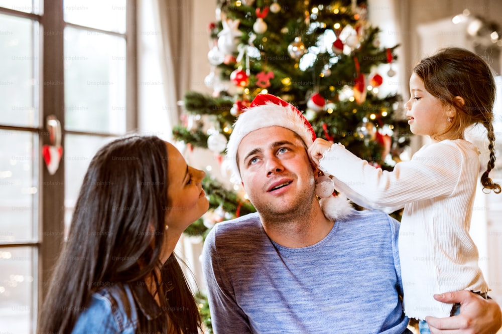 Schöne junge Familie mit kleinem Mädchen, das den Weihnachtsbaum zu Hause schmückt. Tochter gibt dem Weihnachtsmann Weihnachtsschmuck auf die Ohren.