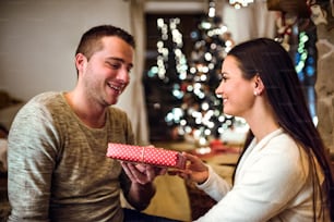 Beautiful young couple sitting on the floor in front of fireplace. Man adn woman giving presents to each other.