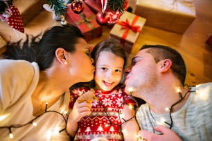 Young parents with their little daughter under Christmas tree. Mother and father kissing their daughter.