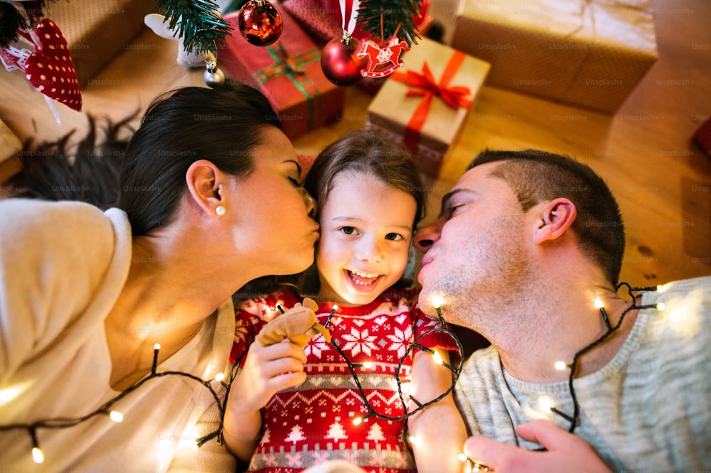 Young parents with their little daughter under Christmas tree. Mother and father kissing their daughter.