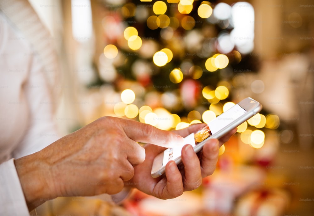 Mujer irreconocible sosteniendo un teléfono inteligente, enviando mensajes de texto, sentada frente al árbol de Navidad iluminado dentro de su casa.
