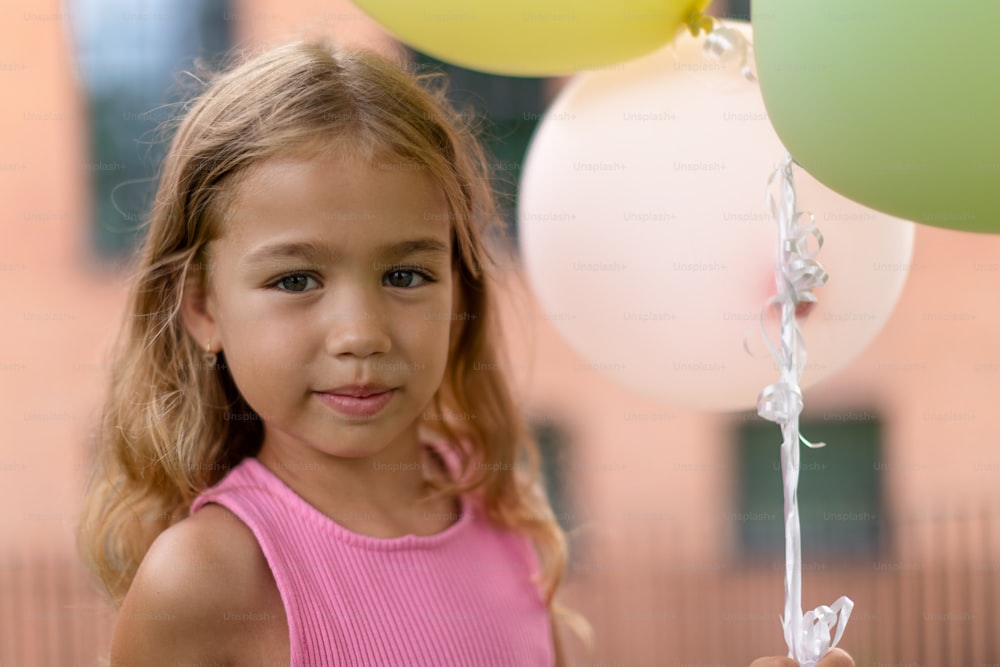 Portrait of little blond girl posing with baloons in city and looking at camera.
