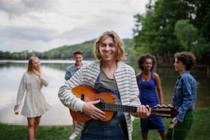 Group of young friends having fun near the lake, laughing and playing guitar.