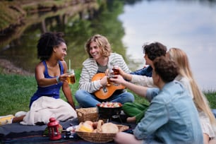 A group of young friends having fun on picnic near a lake, sitting on blanket and toasting with drinks.